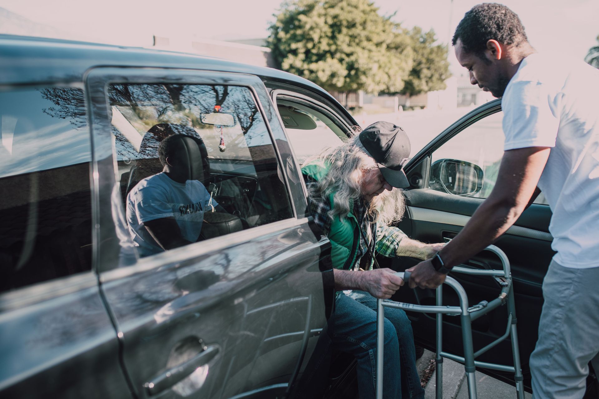 Man loading a woman into a wheelchair beside a car, with a child seated inside, outdoors in daylight