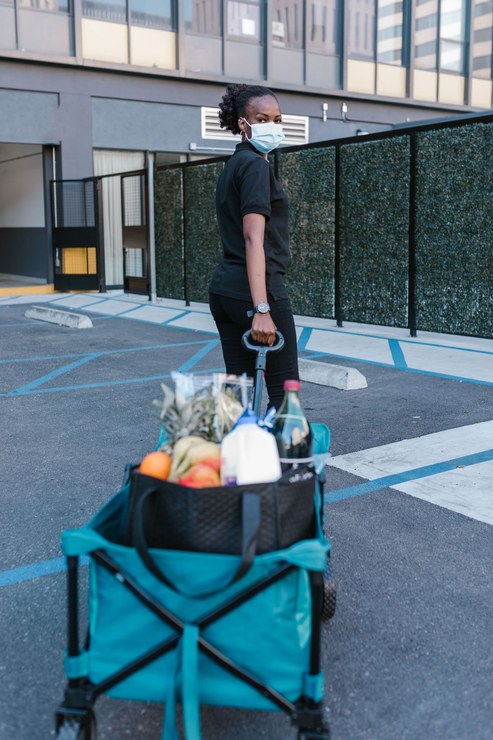 Person wearing a mask pushes a teal cart loaded with groceries across a parking lot.