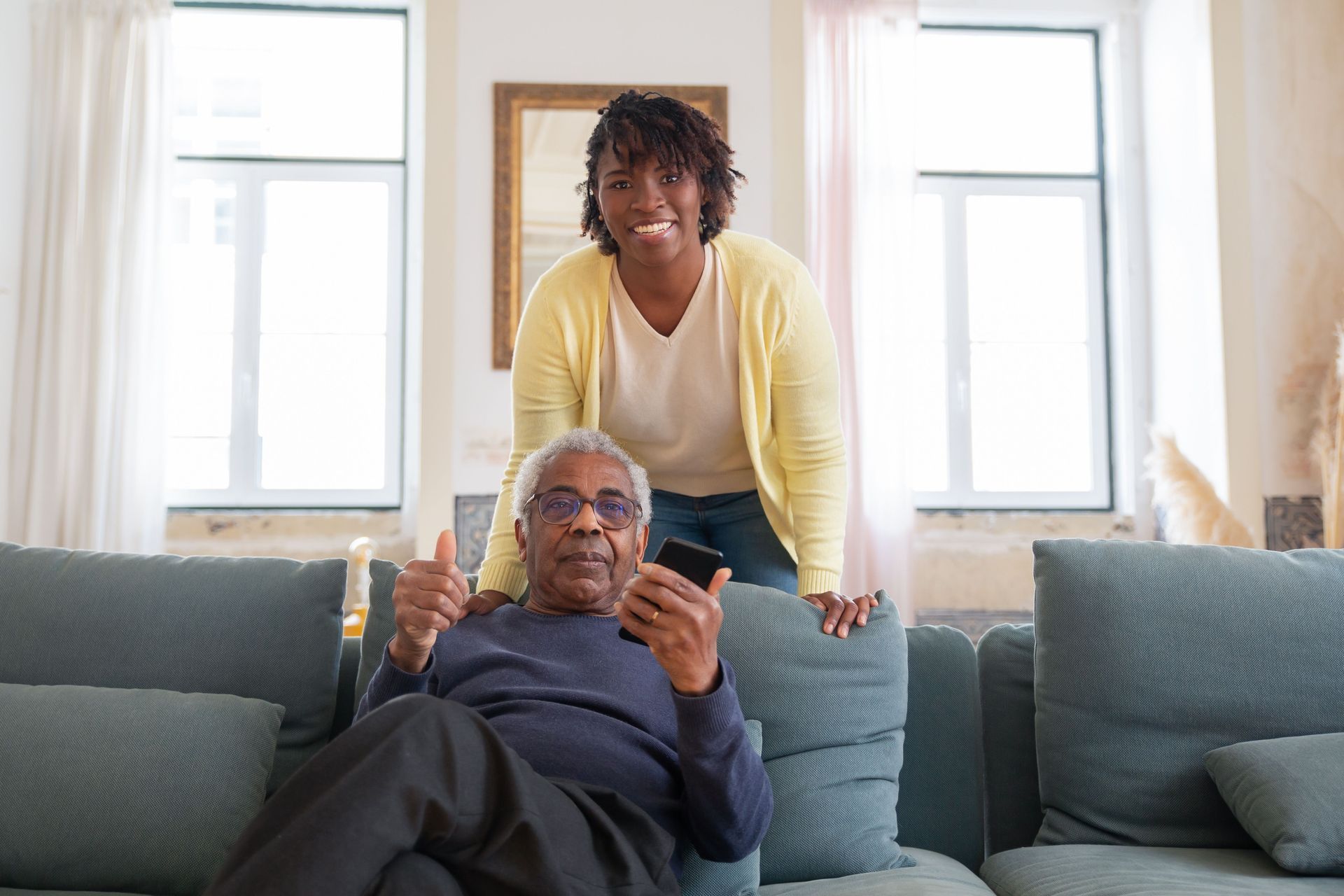 Older man reclining on a couch as a woman stands behind him, smiling in a bright living room