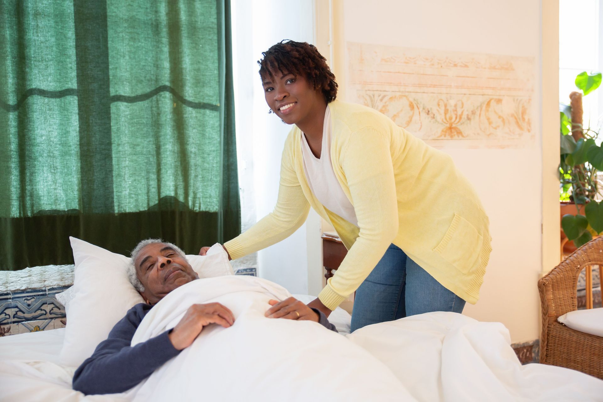 Caregiver leaning over a patient in bed, adjusting white bedding in a bright bedroom.