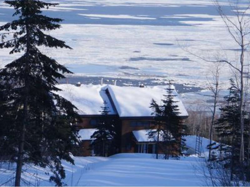 Snow-covered house in a winter landscape with frozen water in the background and evergreen trees.