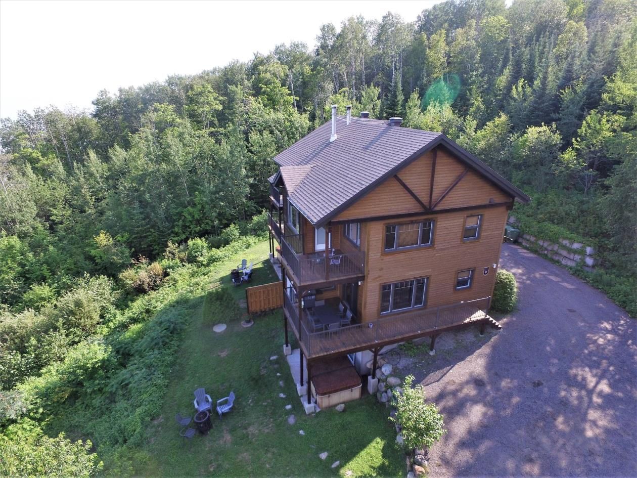 Wooden house nestled in a green forest; has a deck and driveway.