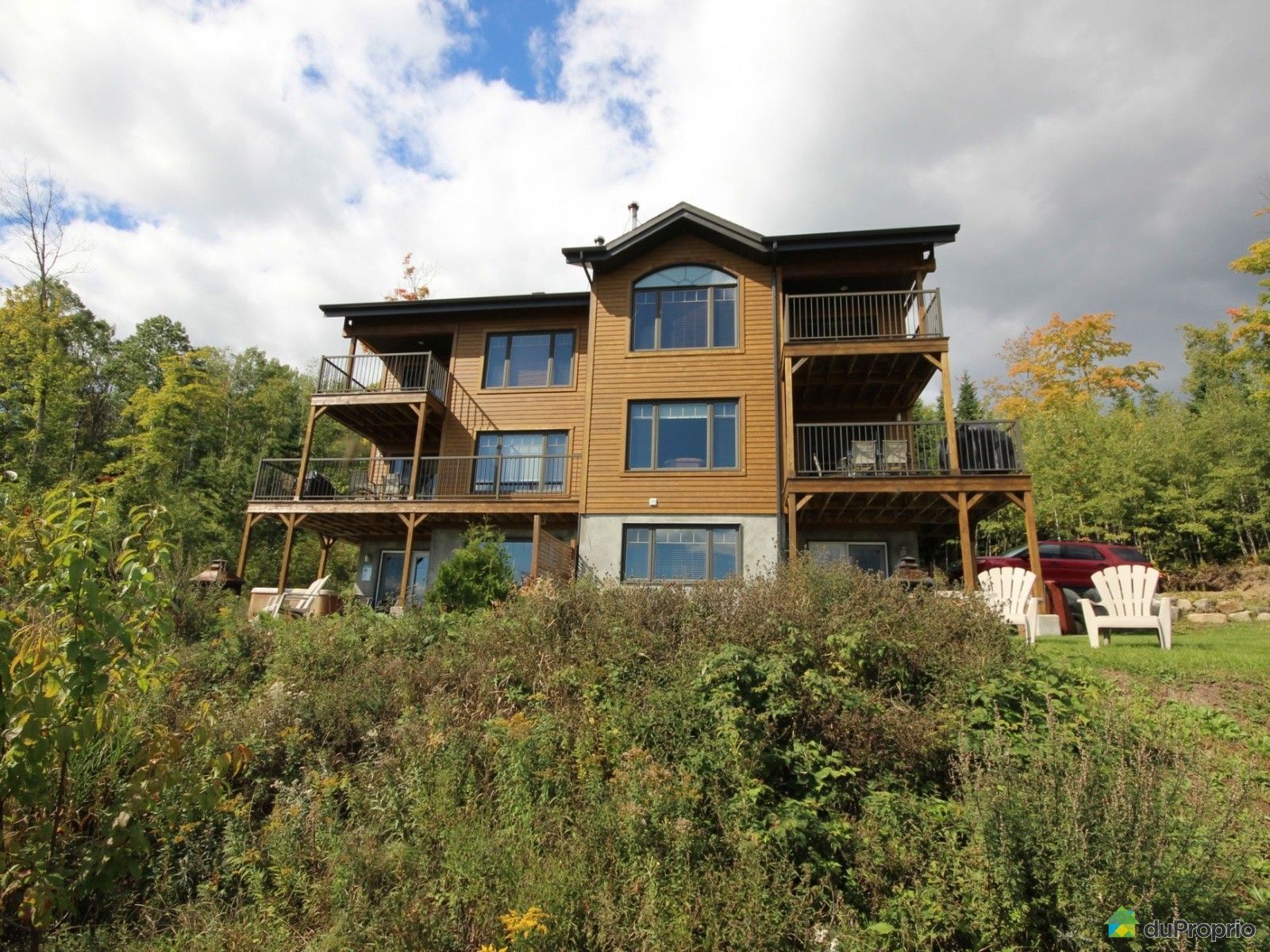 Two-story wooden house with balconies, windows, and decks, set amidst foliage under a cloudy sky.