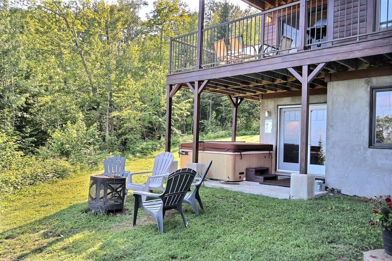 A hot tub and seating area under a deck of a house, surrounded by trees.