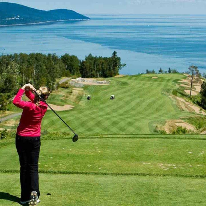 Woman in red golf shirt swings on a green, ocean and mountains in the background.