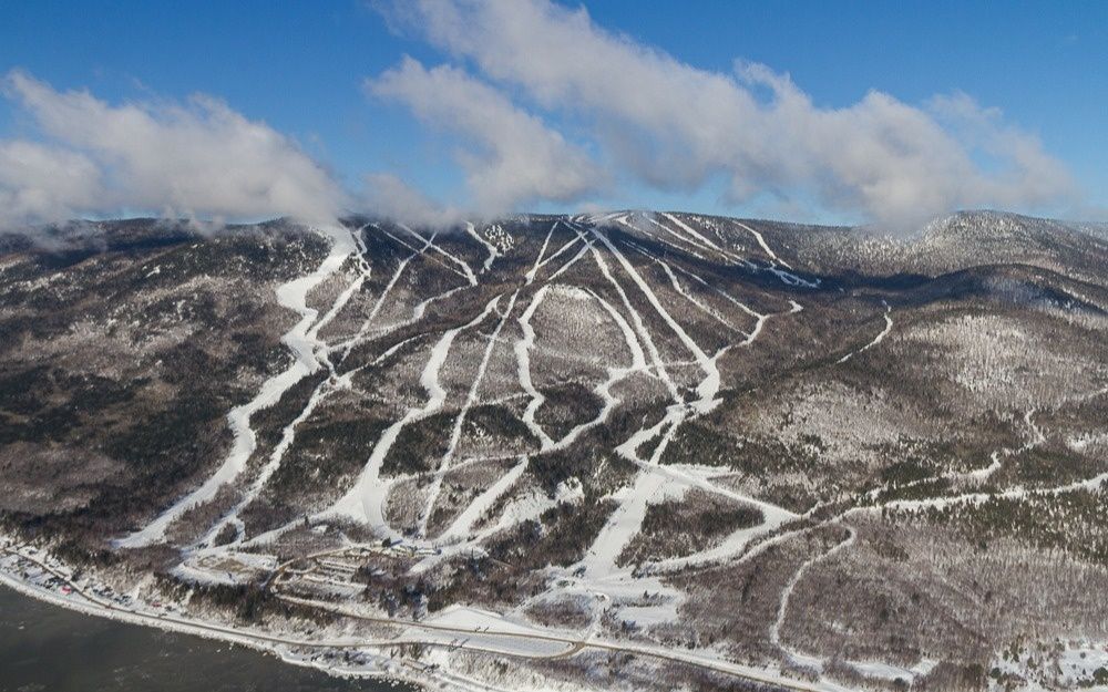 Montagne enneigée avec des pistes de ski tracées à travers les arbres. Ciel bleu parsemé de nuages ​​blancs.