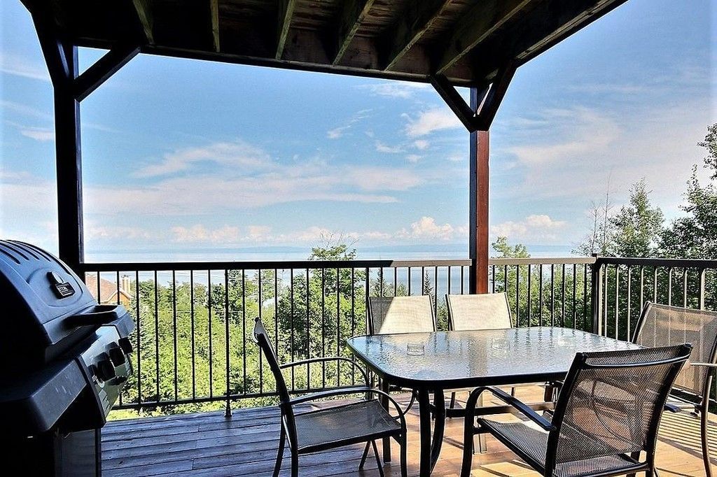 Deck with grill, table, chairs, and railing overlooking a body of water and trees under a blue sky.
