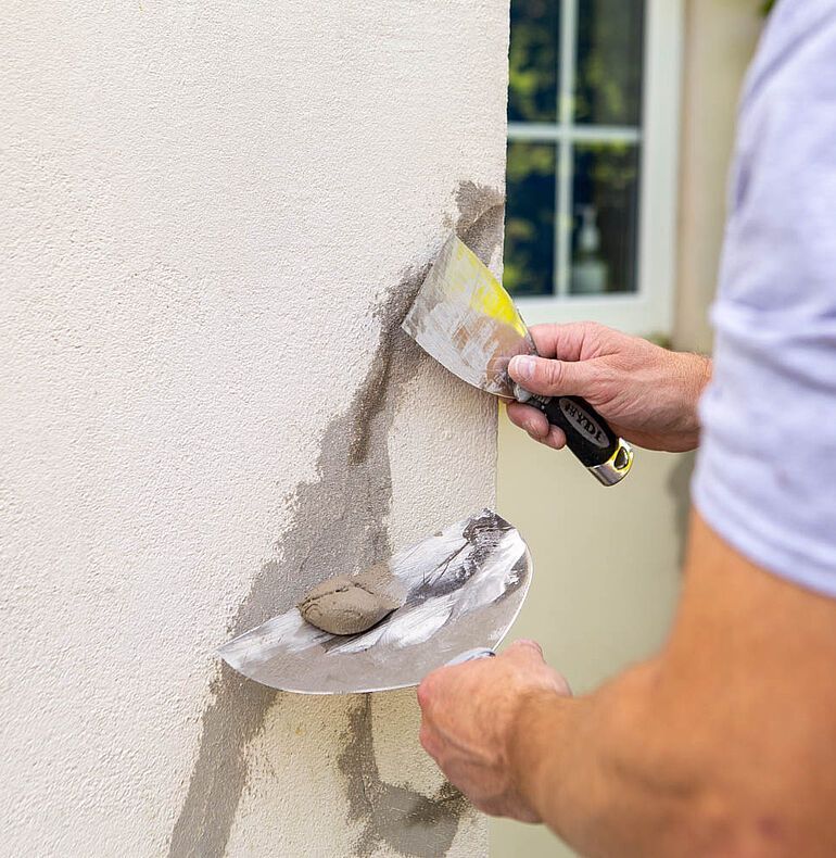 Person using a trowel to apply stucco to an exterior wall near a window.