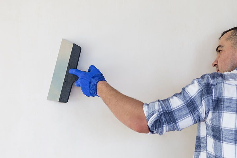 Person with blue glove uses a putty knife to smooth plaster on a white wall.