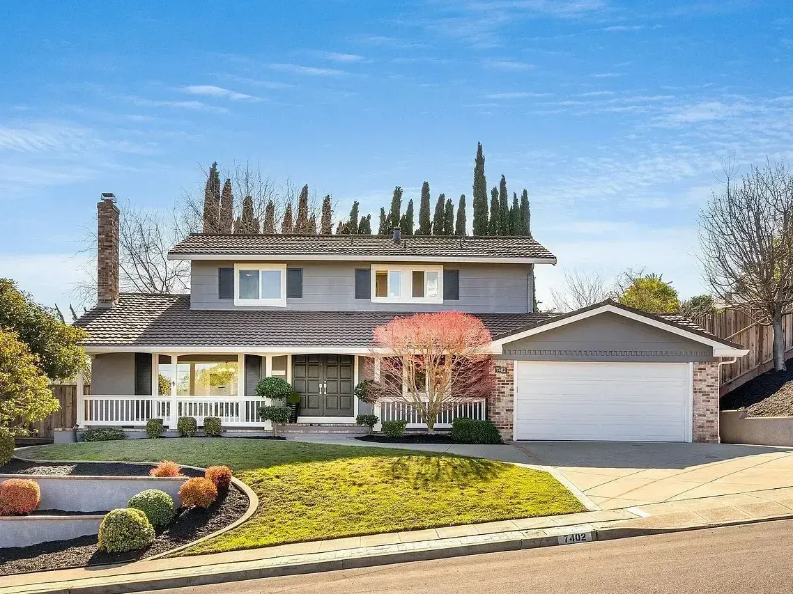 Two-story gray house with white trim, garage, and landscaped yard under a blue sky.