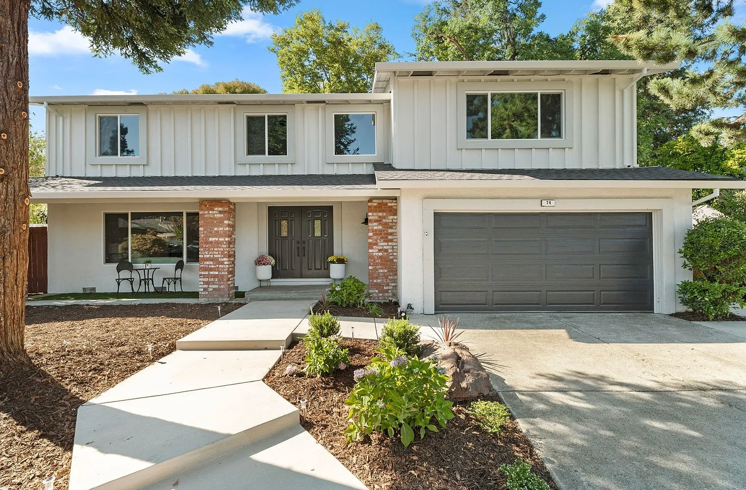 Two-story light gray house with dark gray garage door, red brick accents, and a paved walkway leading to the front door.