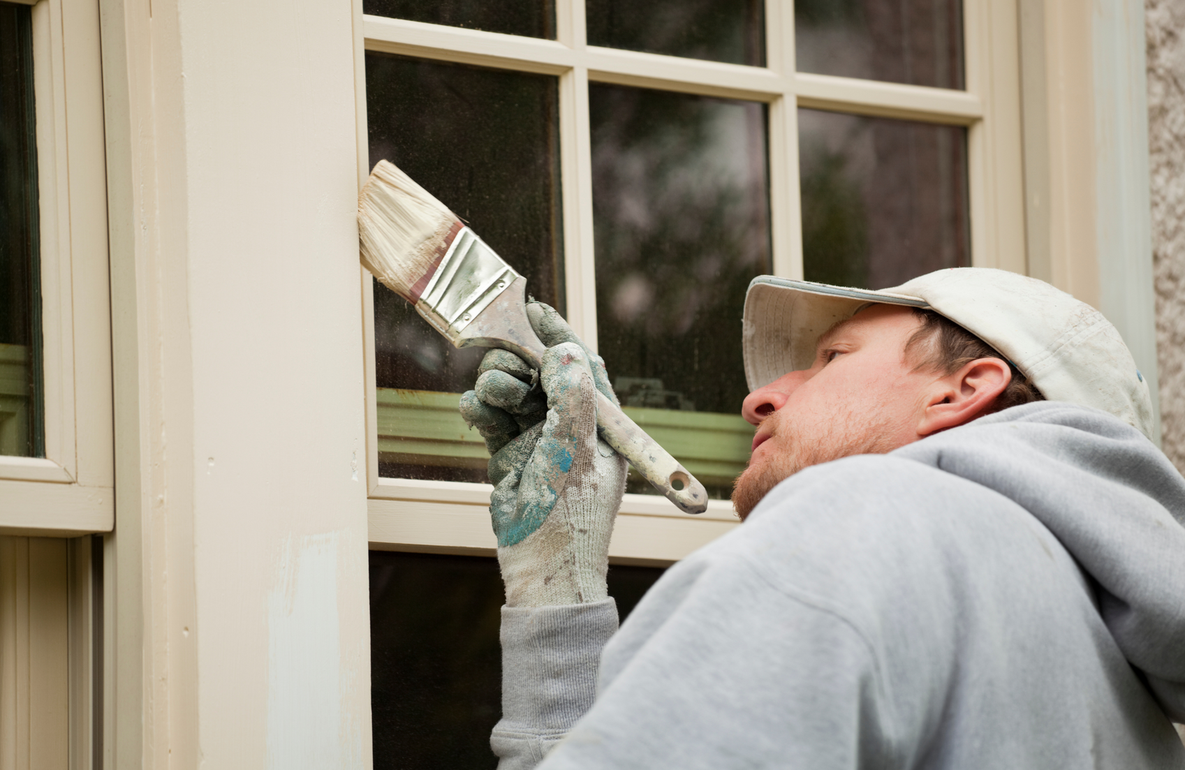 Person painting window frame with brush. Outdoors, light colors.