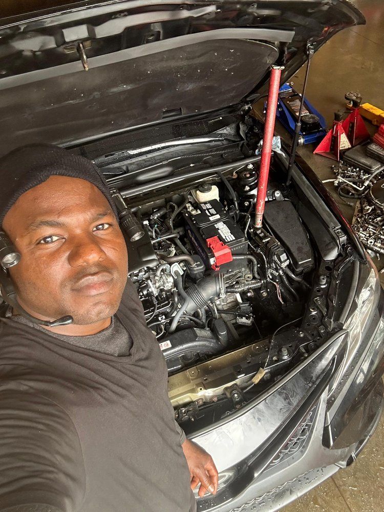 Mechanic working under an open car hood in a repair shop, with tools and engine parts visible.
