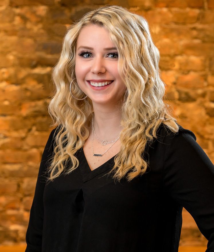 Woman with curly blonde hair smiles at the camera in front of a brick wall.