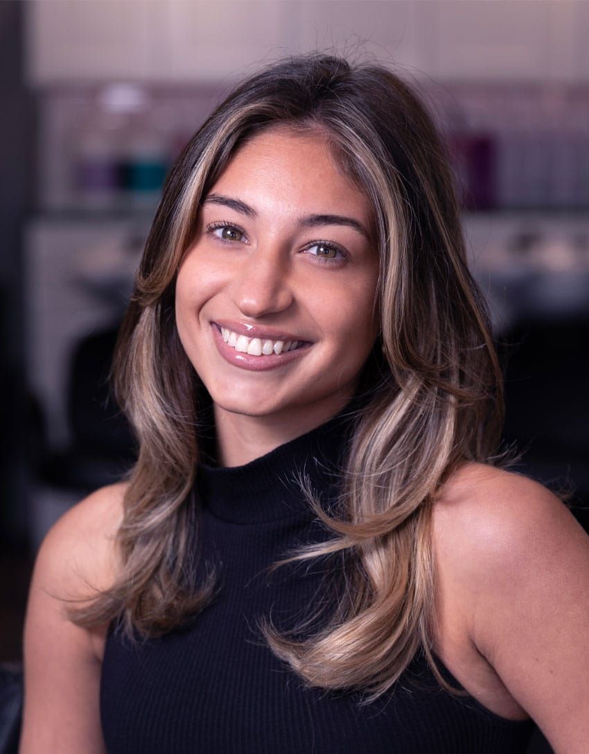 Woman smiling, shoulder-length brown hair with blonde highlights, wearing a black top. Salon background.