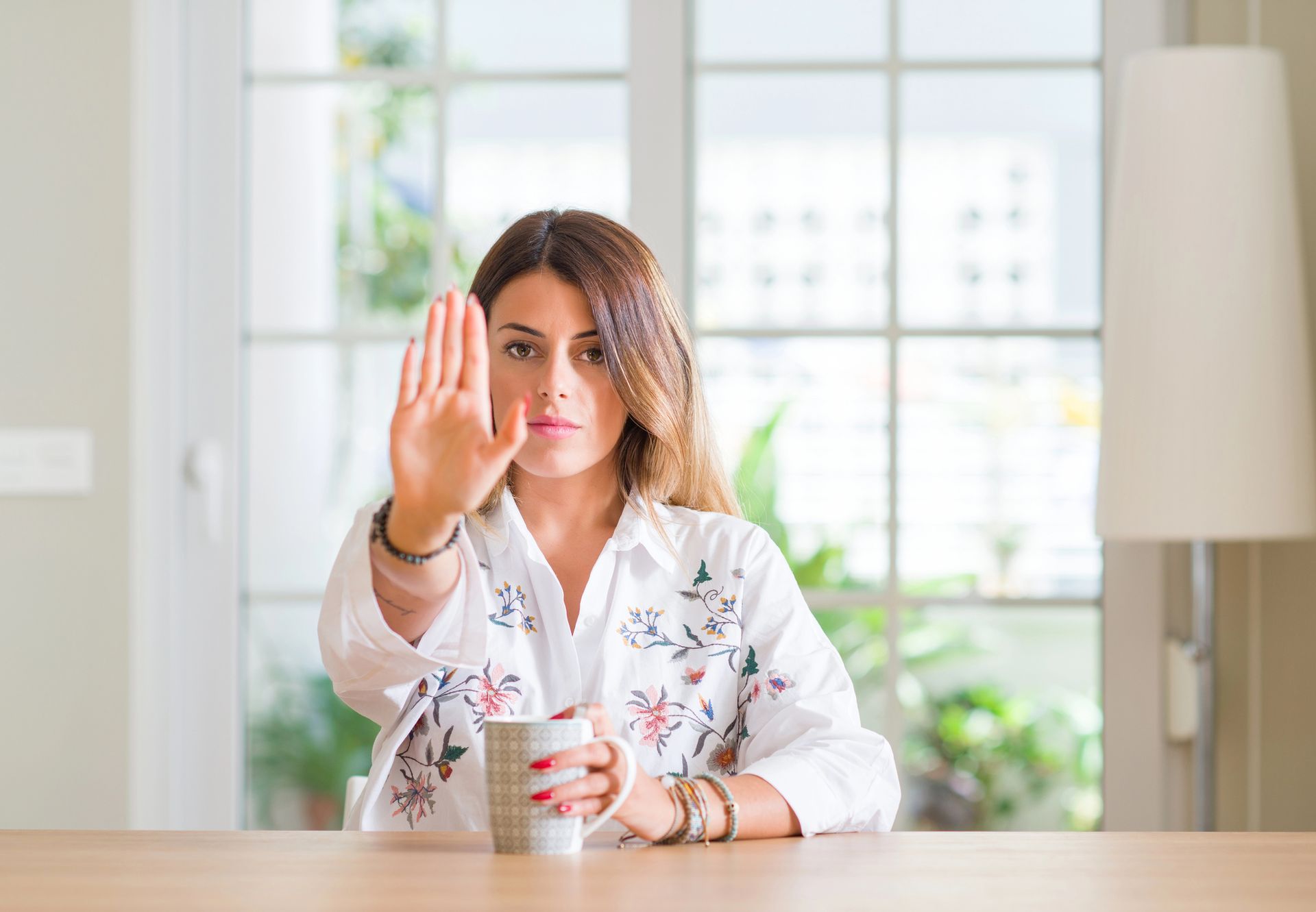 A woman is sitting at a table holding a cup of coffee and making a stop sign.