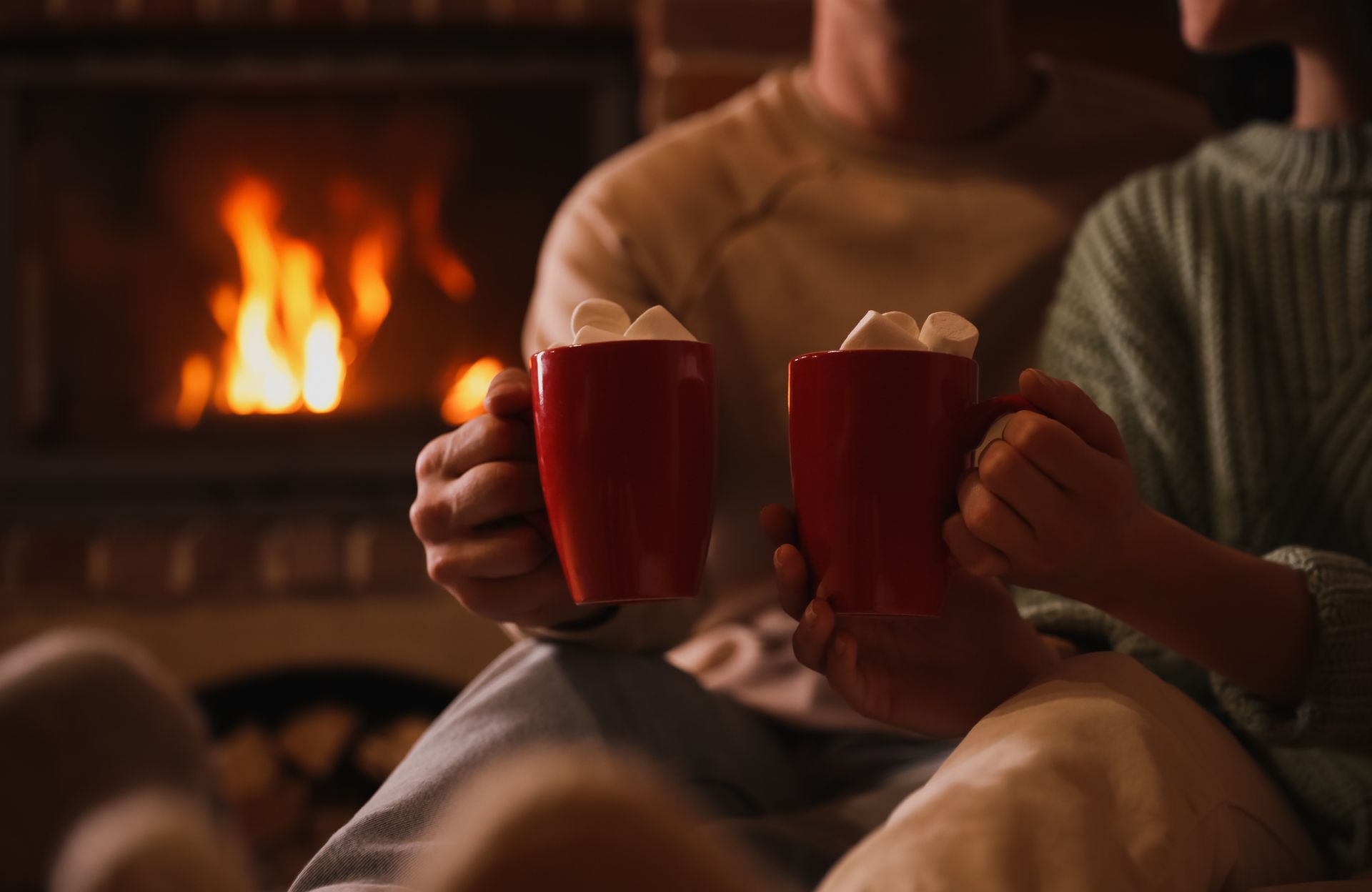 Couple holding red mugs with marshmallows in front of a fireplace.
