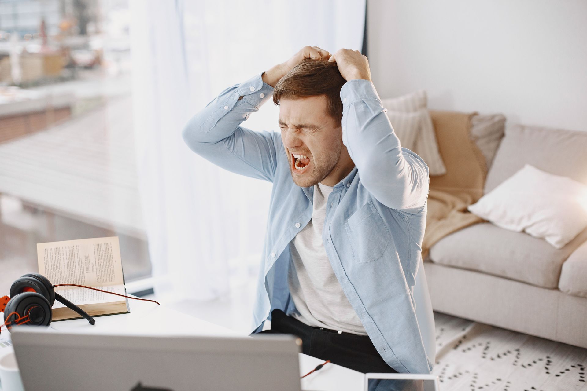 Stressed man holding his head at a laptop on a bright desk with coffee and papers nearby