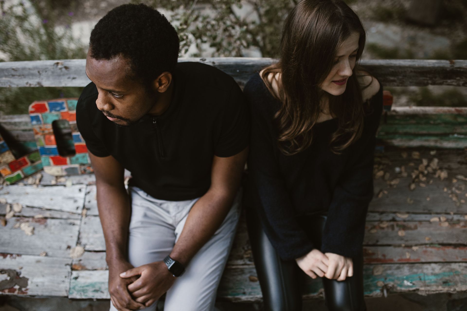 Two people sit on a weathered bench, leaning forward in a quiet outdoor setting.