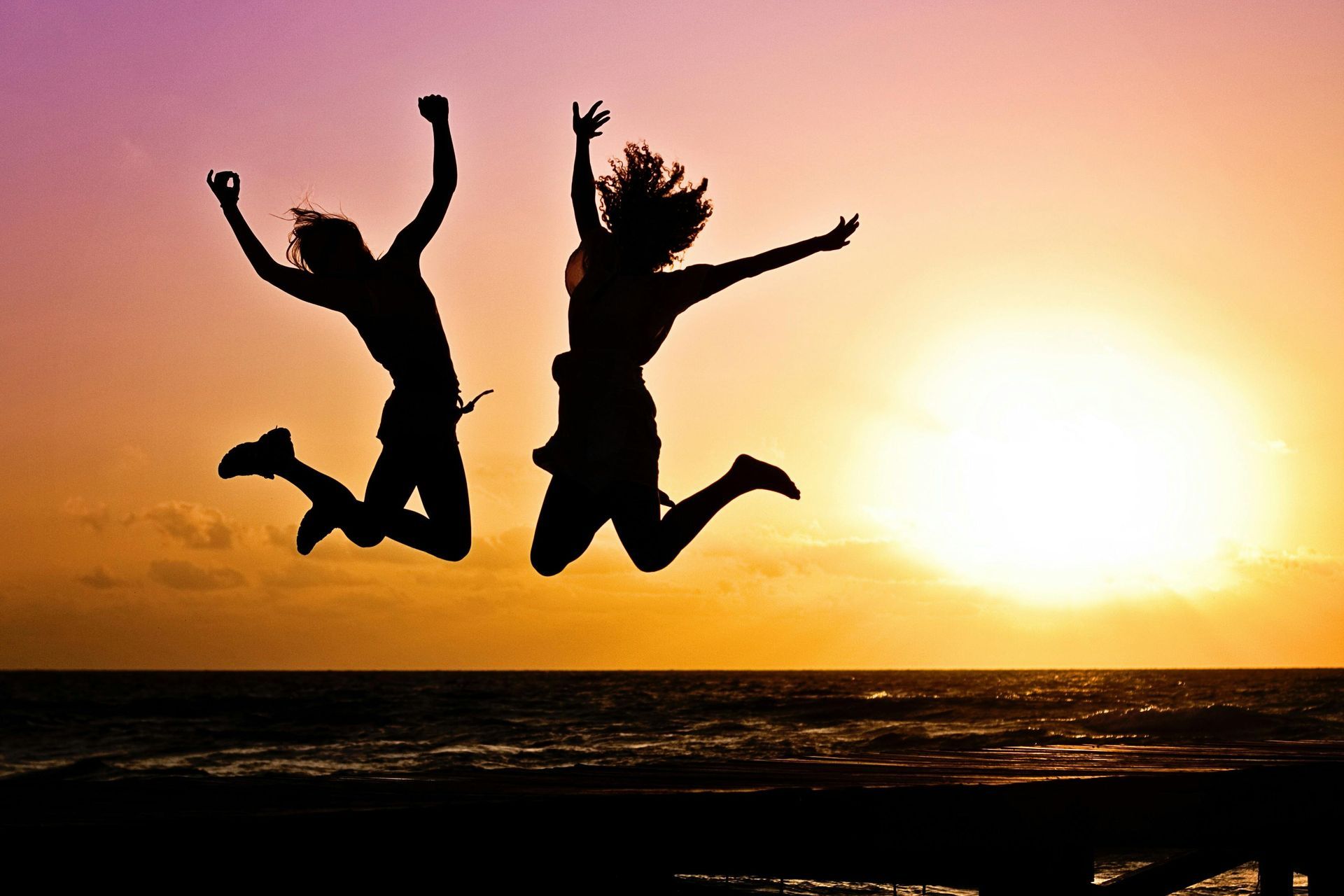 Two people silhouetted jumping in air, arms raised, at sunset over the ocean.