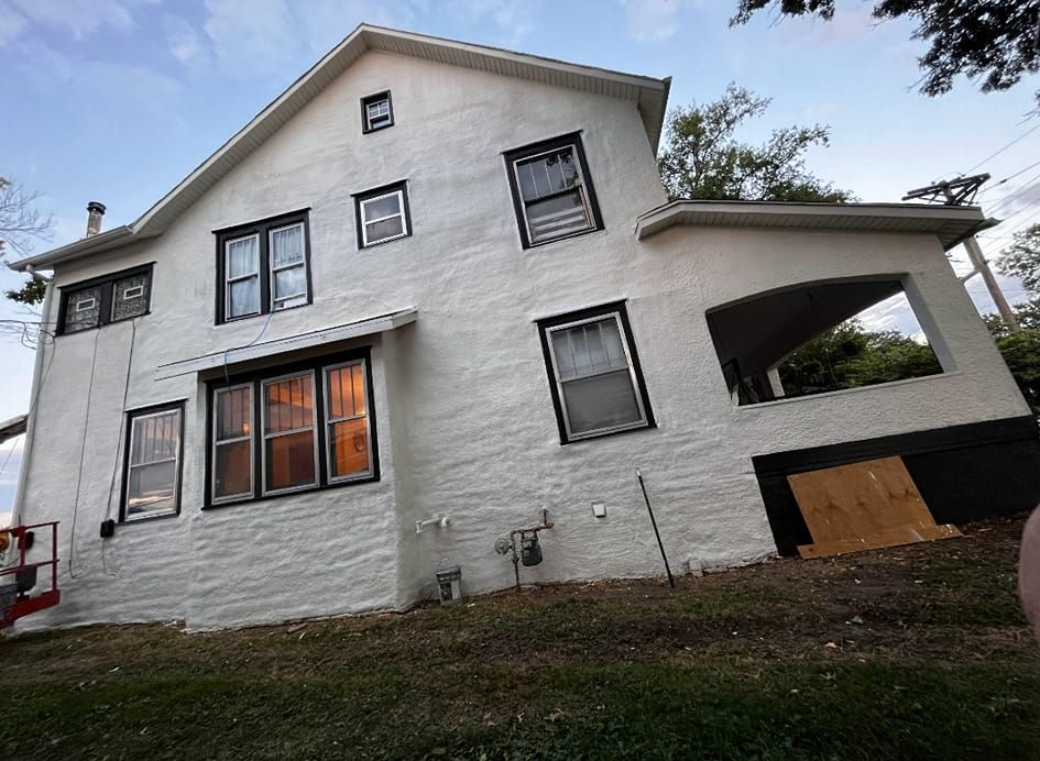 White two-story house with missing siding and a porch. Green grass and blue sky.