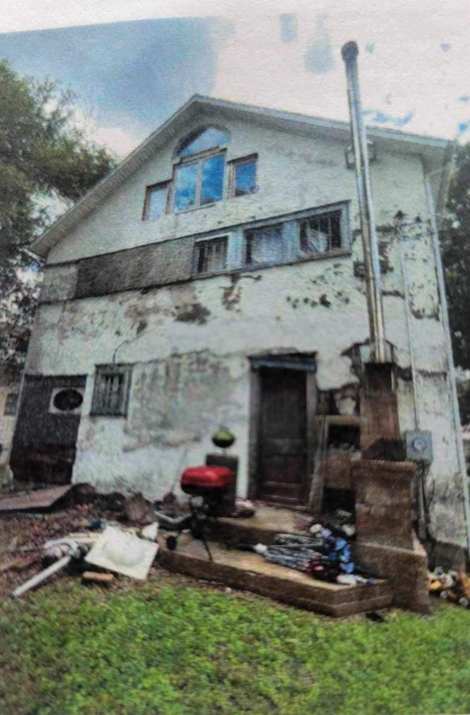 Weathered two-story house with peeling paint and debris on the lawn. A chimney rises beside the building.