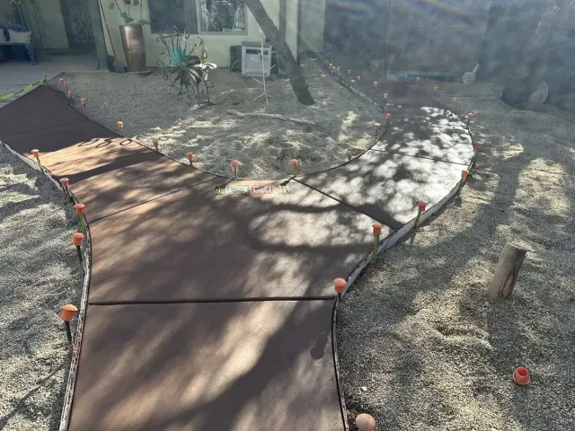 Brown concrete pathway with curved edge, surrounded by gravel, marked by orange leveling pegs.