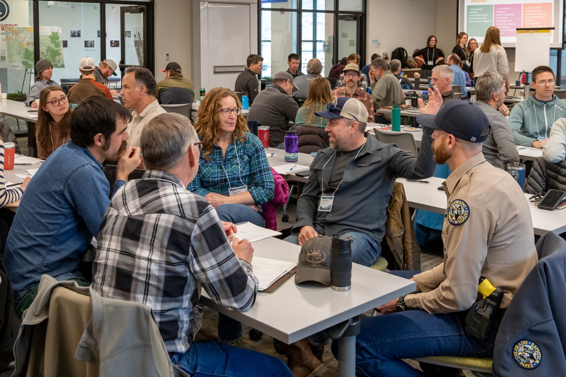A group of people sit at tables in a brightly lit room, engaged in conversation during a workshop or conference in roaring fork colorado