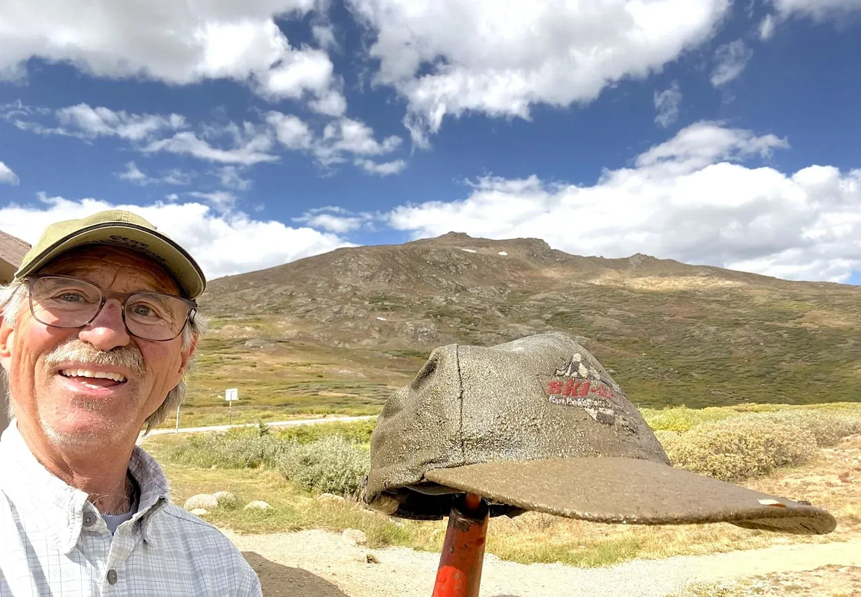 A smiling person wears glasses and a hat while holding up an old, weathered baseball cap against a mountain landscape in colorado