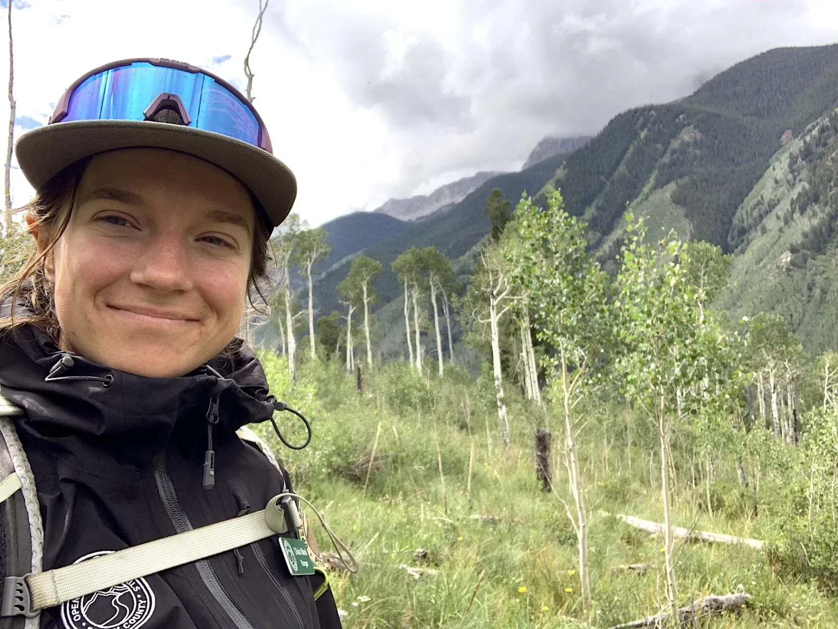 A smiling hiker wearing a cap and jacket stands in a lush, green mountain meadow with aspen trees and rugged peaks.