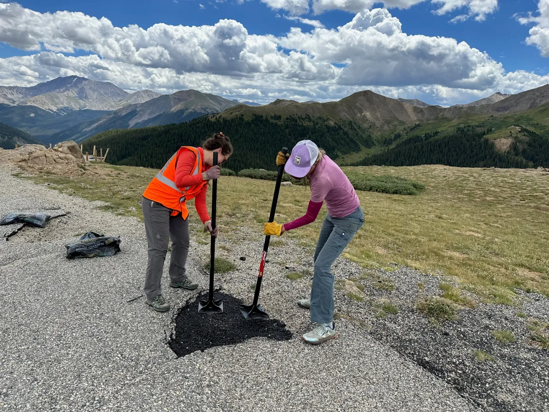 Two people use hand tools to work on a rocky, mountain-side path under a cloudy sky in the roaring fork watershed
