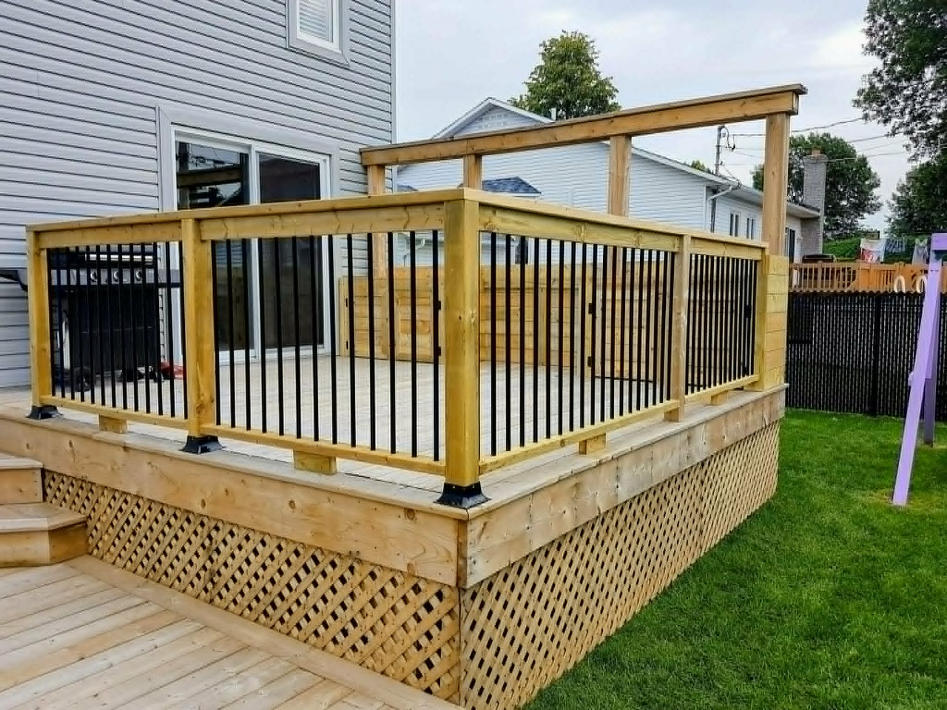 Terrasse en bois avec rambarde et plinthe en treillis, donnant sur une maison et une pelouse verdoyante.
