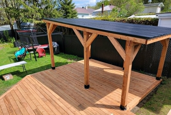 Terrasse en bois à l'arrière de la maison, avec une pergola au toit incliné et des panneaux solaires noirs, à côté d'une pelouse.