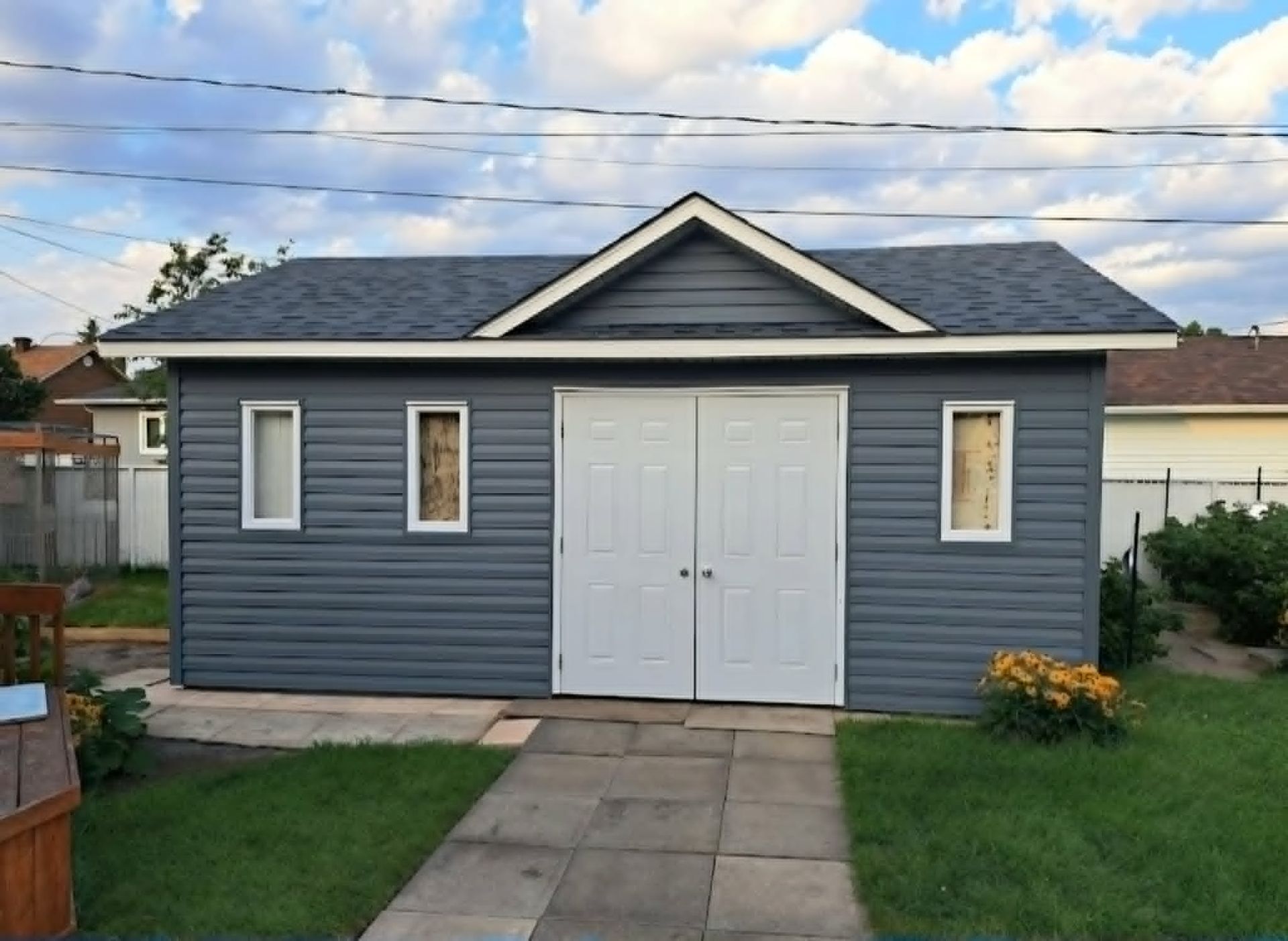 Petit abri de jardin bleu avec double porte blanche et toit gris, derrière une terrasse en béton.