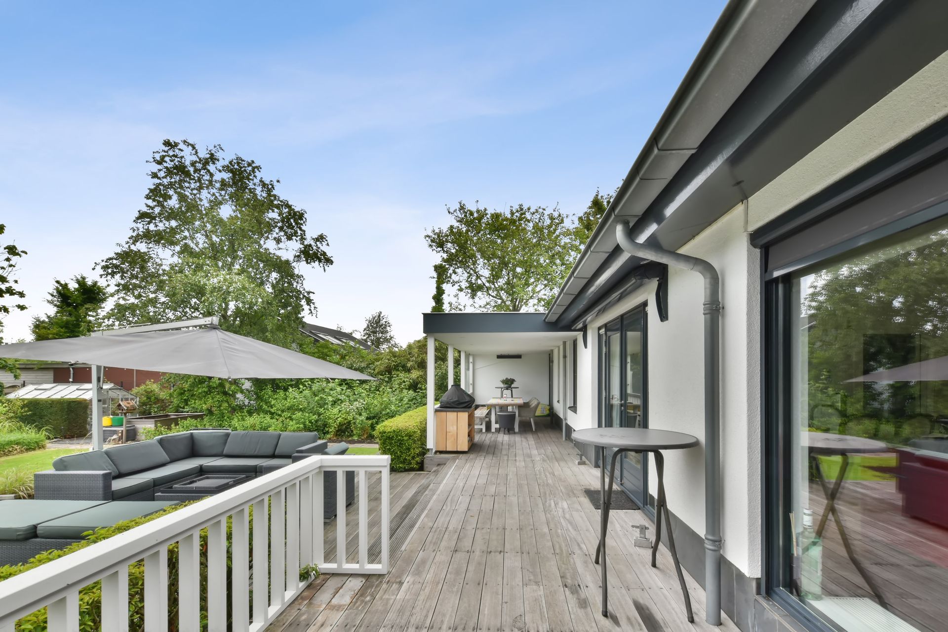 Spacieuse terrasse en bois jouxtant une maison moderne, avec mobilier de jardin, parasol et arbres sous un ciel bleu.
