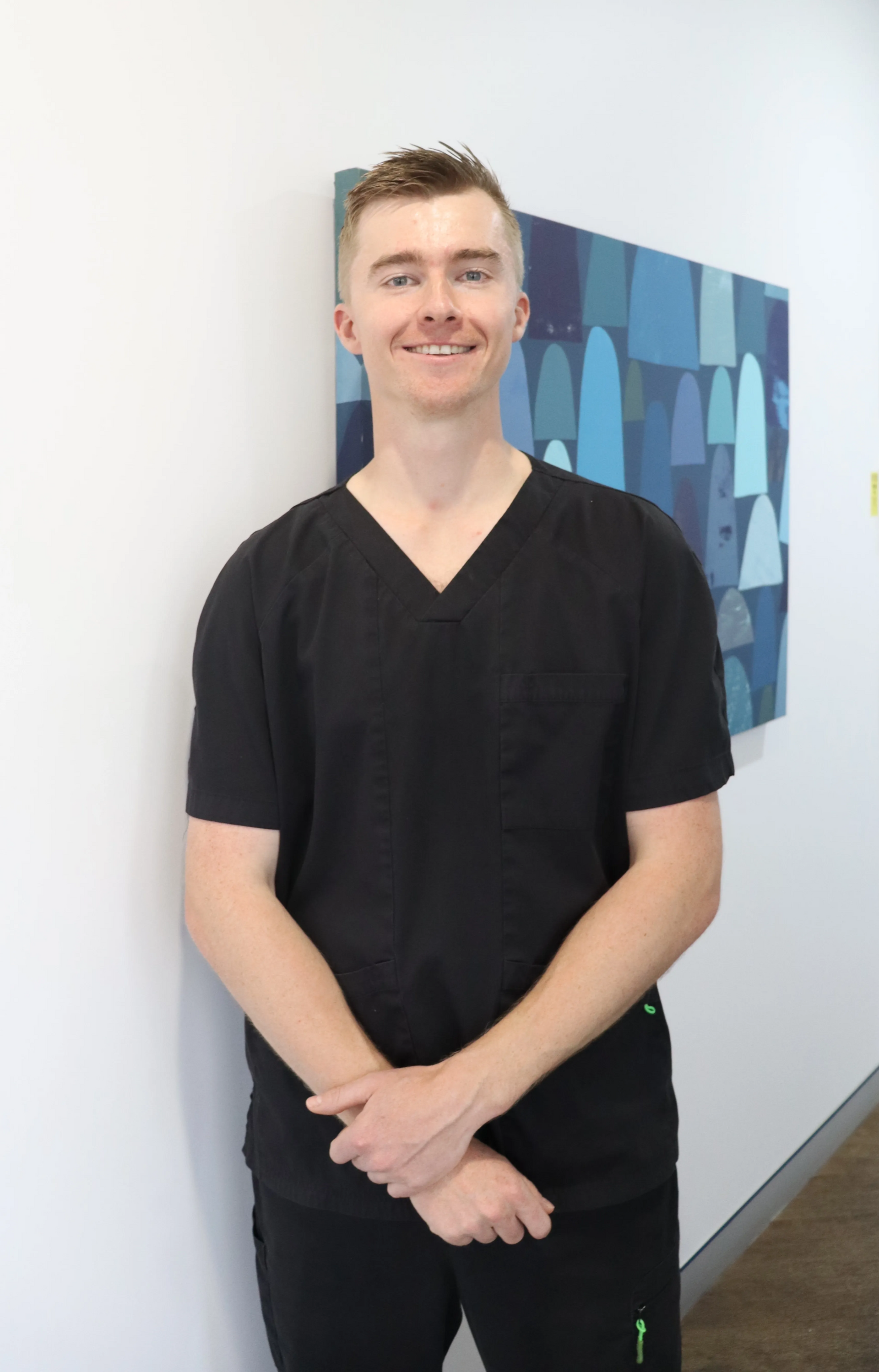Man Standing Indoors In Black Scrubs, Smiling With Hands Clasped, Beside A Blue Abstract Wall Painting — McDonald Dental in Gympie, QLD