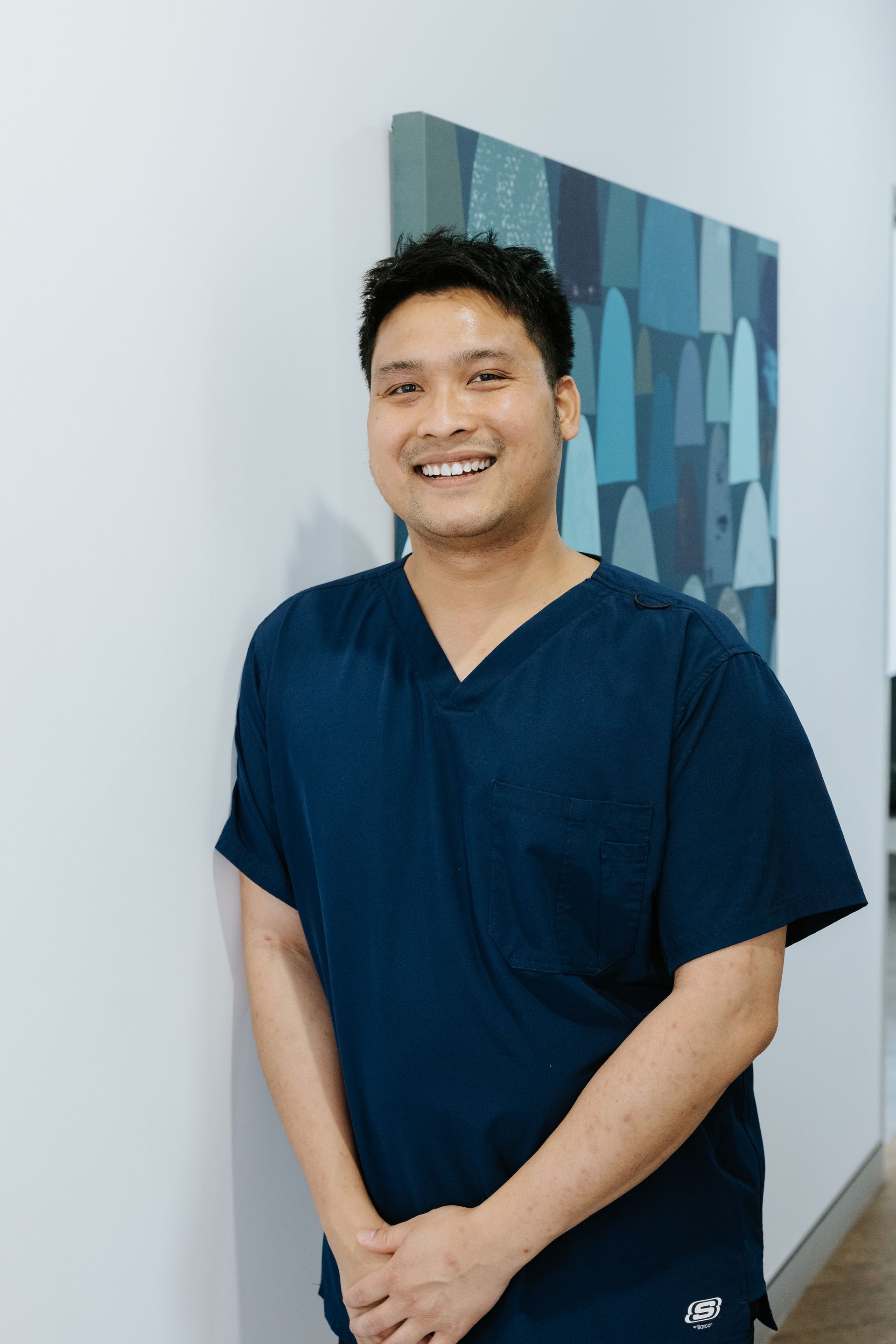 Smiling Person In Navy Scrubs Standing Indoors Beside A Wall And Framed Artwork — McDonald Dental in Gympie, QLD