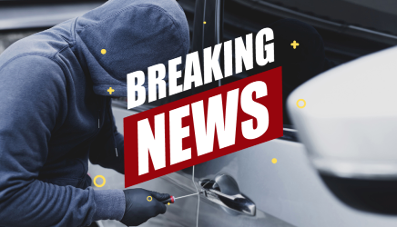 A man in a hoodie is breaking into a car. A sign stating 