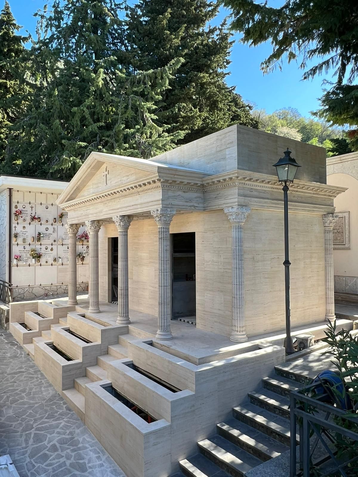 Piccolo mausoleo in pietra bianca con scalini e colonne in un cimitero, circondato da alberi ad alto fusto.