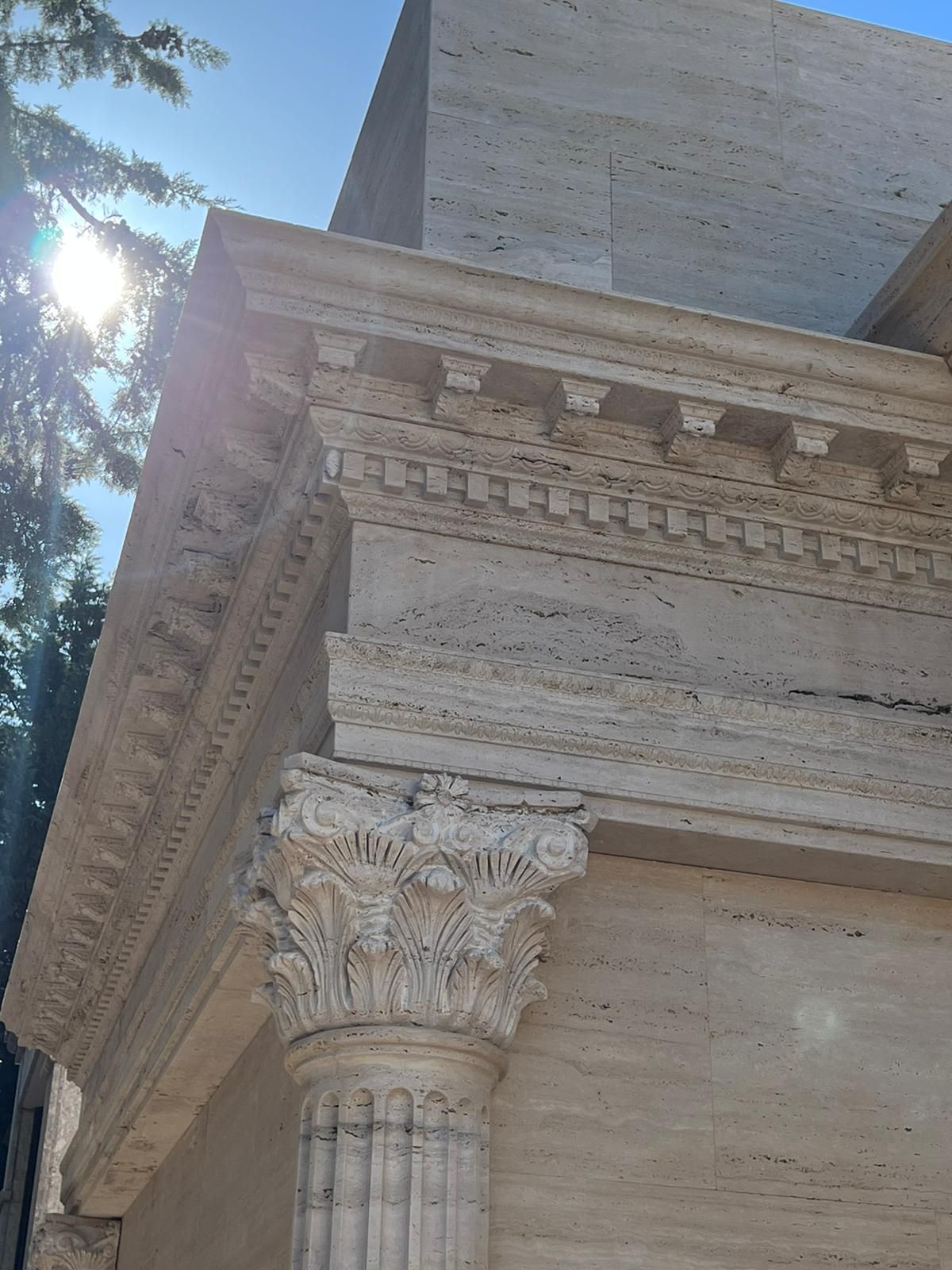 Angolo di un edificio in pietra con colonna e cornicione decorati, illuminato dalla luce del sole; alberi visibili a sinistra.