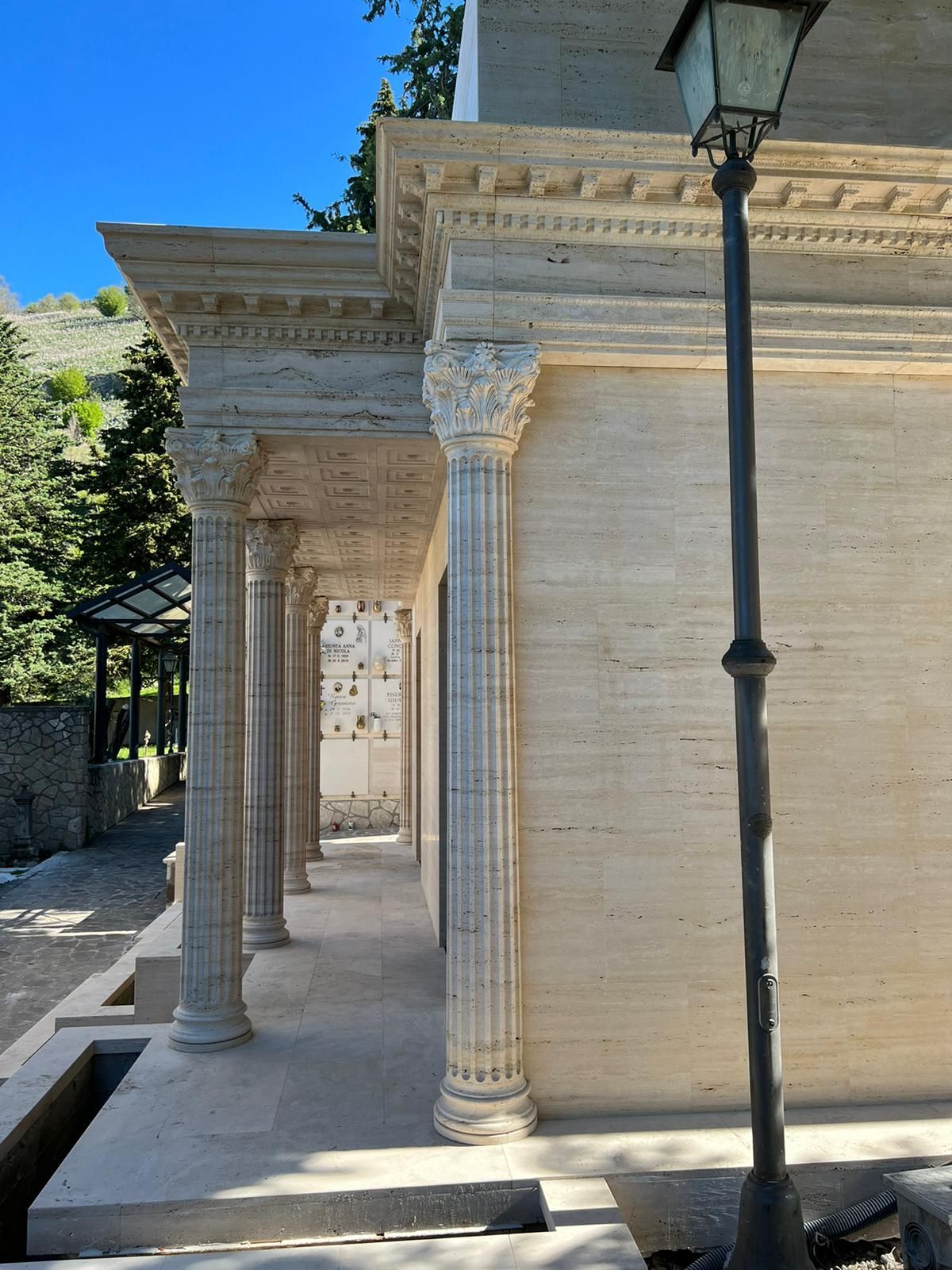 Un colonnato in pietra costeggia un edificio di colore chiaro, con un lampione nero in primo piano.