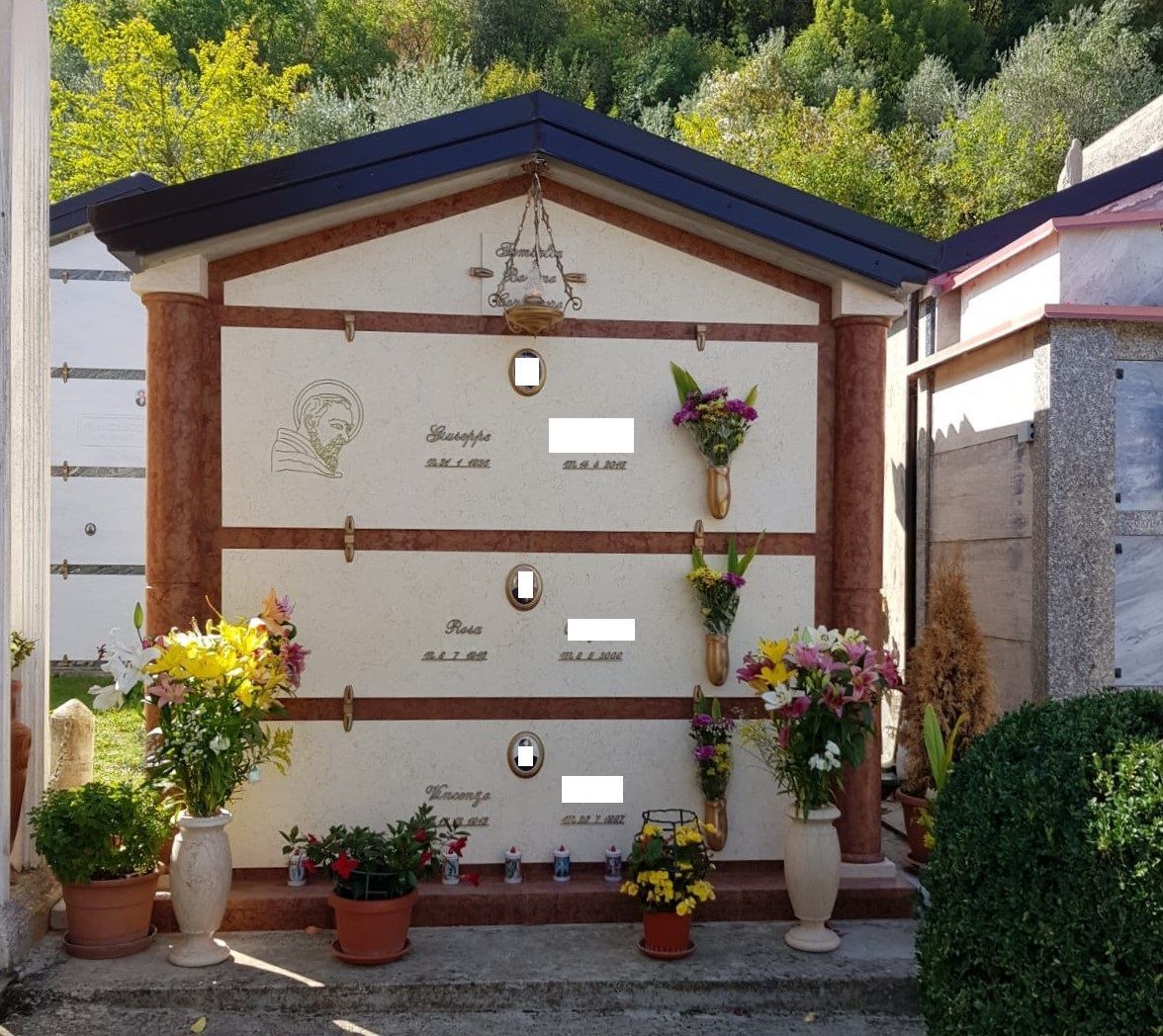 Nicchia cimiteriale decorata con fiori, lapidi e vasi, incastonata in un pendio boscoso.
