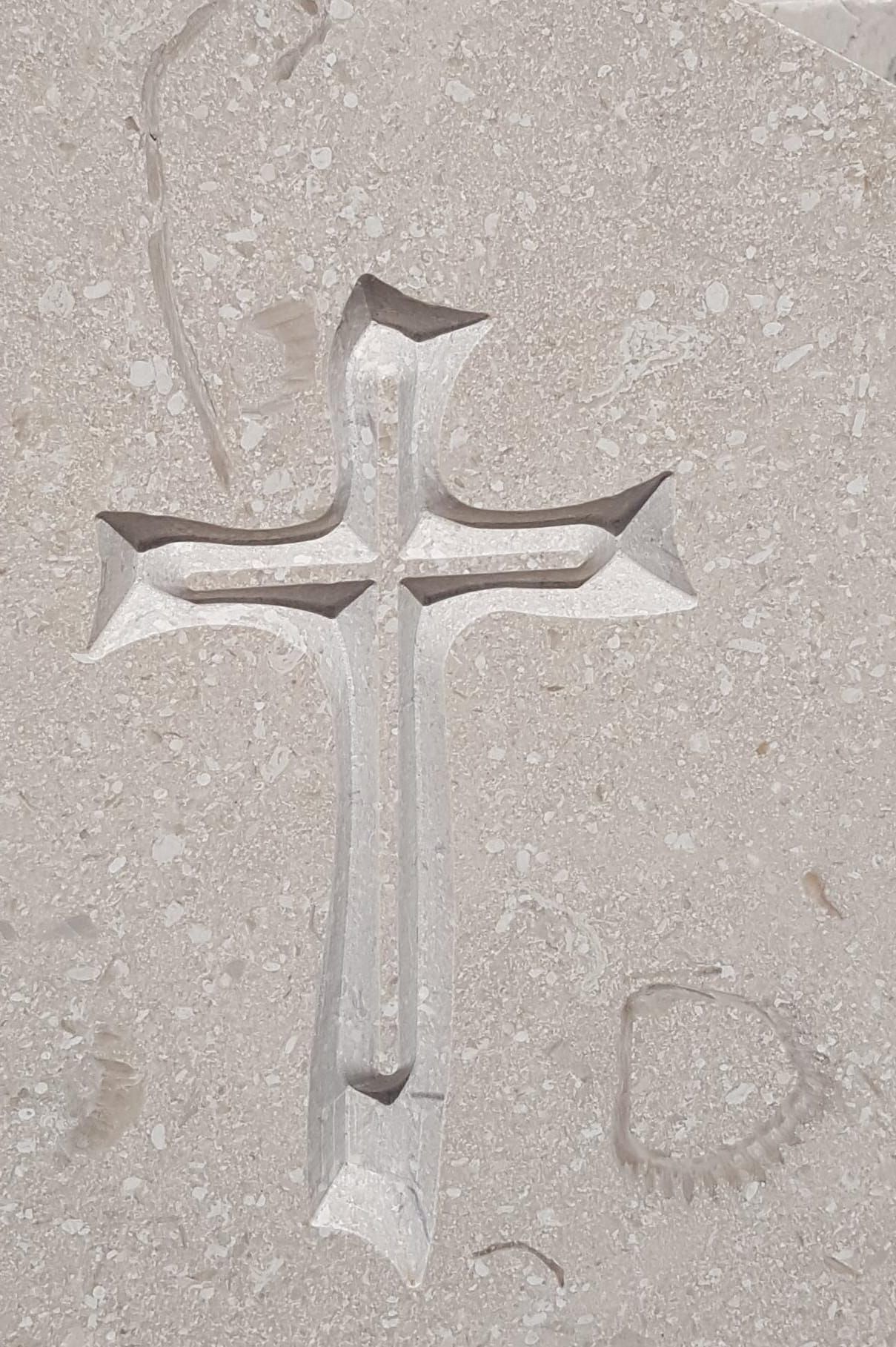 Croce scolpita in pietra chiara con una piccola forma a fiamma nella parte superiore centrale.