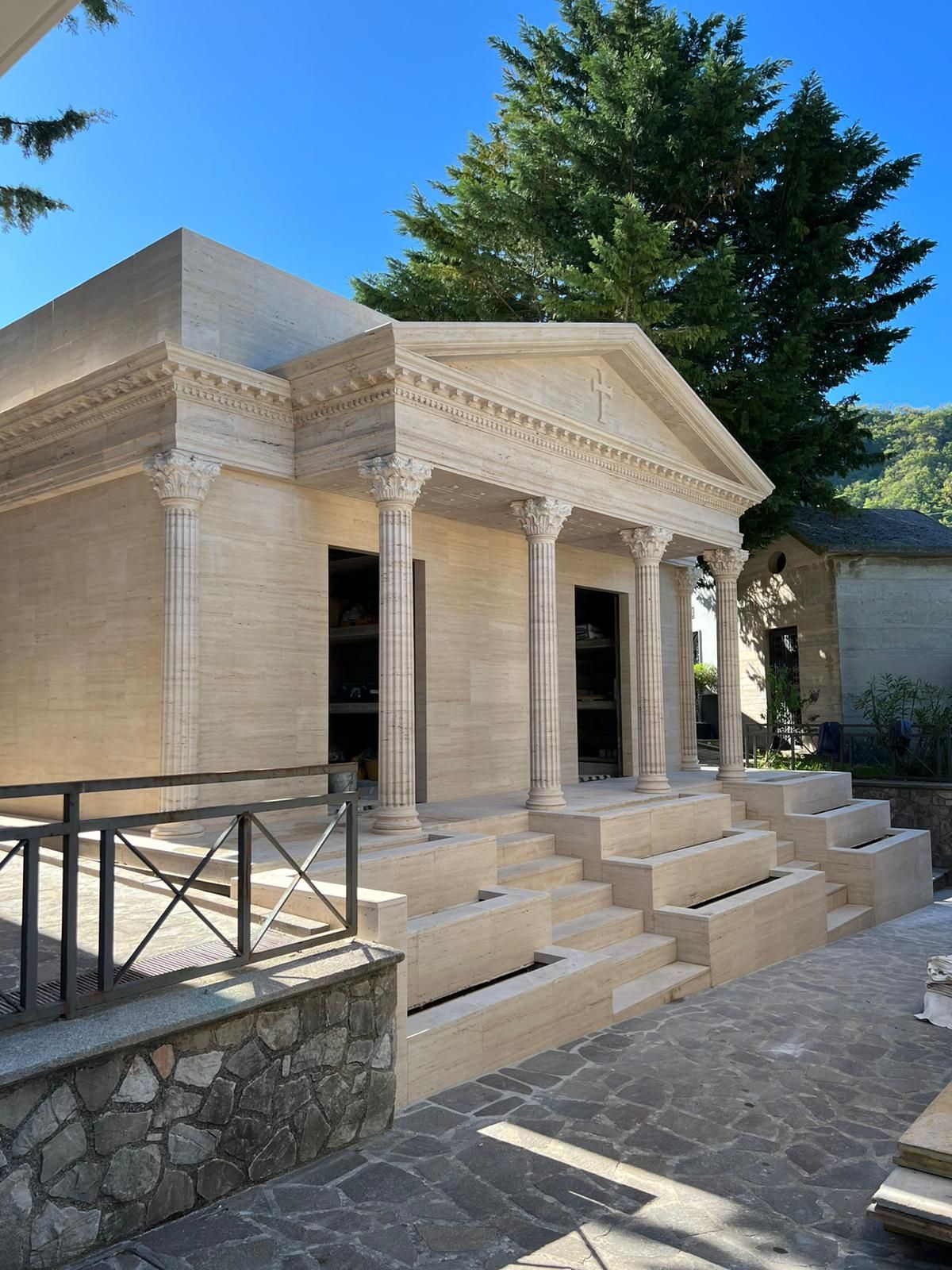 Facciata del tempio in pietra con colonne e scalini, incastonata in un cortile soleggiato con alberi e un cielo azzurro.