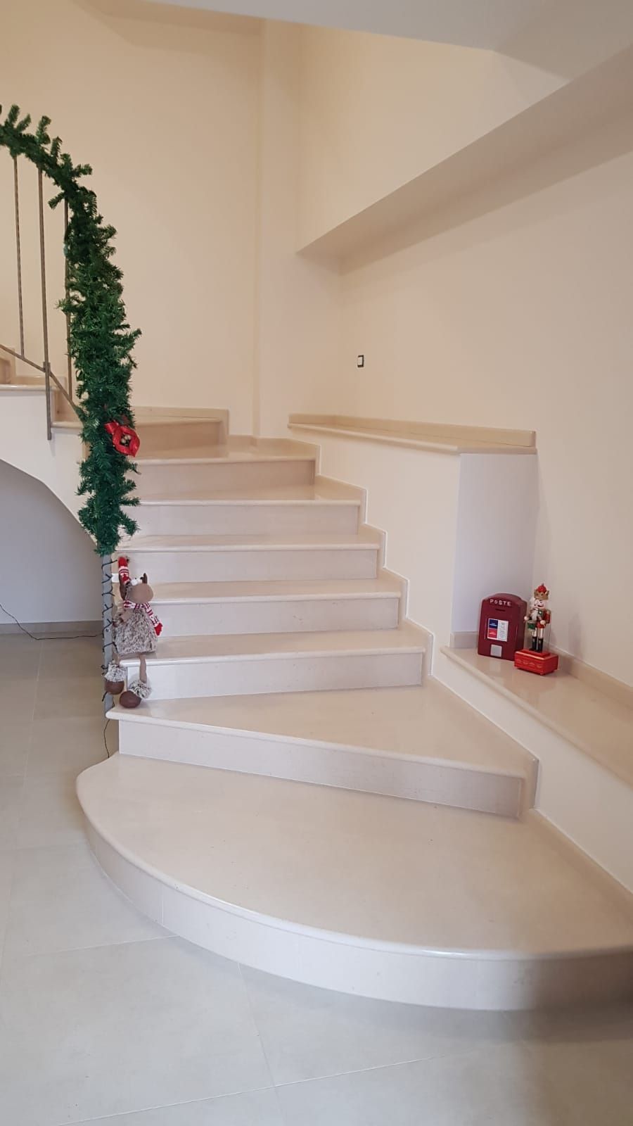 Scala curva bianca con pareti beige e una ghirlanda natalizia sulla ringhiera, oltre a piccole decorazioni sul pianerottolo.