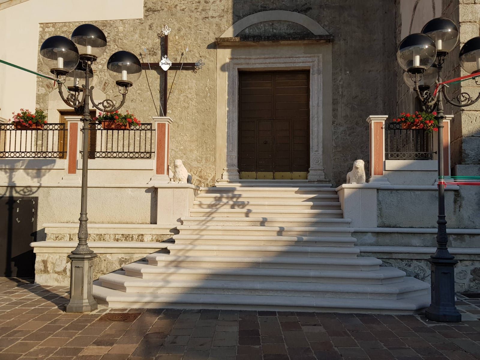 Una scala in pietra conduce a una porta di legno su un edificio beige illuminato dal sole, con lampioni e ringhiere decorate.
