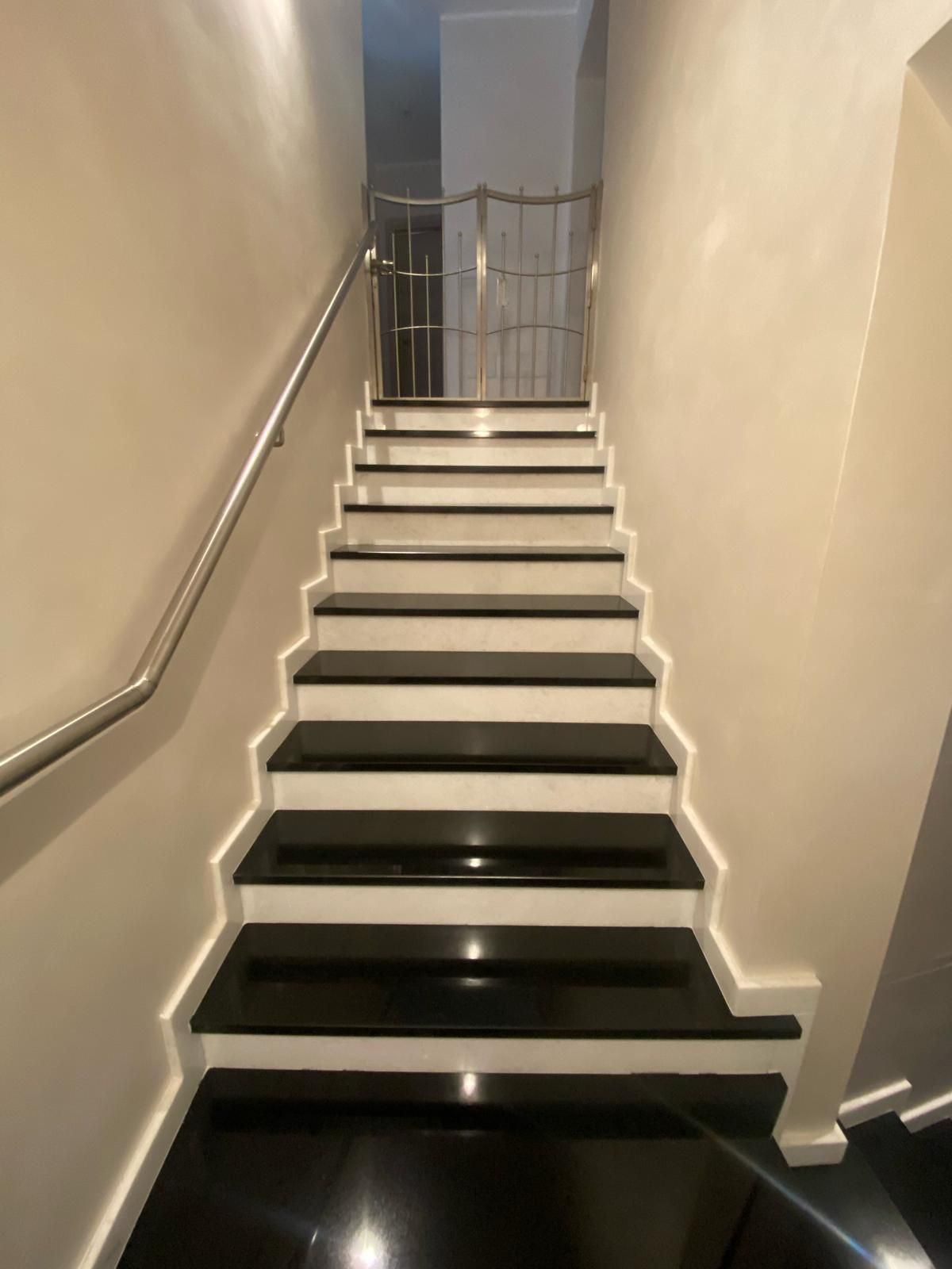 Scala interna stretta con pareti beige, gradini neri e corrimano in metallo che sale verso l'alto.