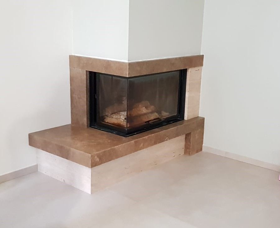 Camino ad angolo con cornice in pietra beige e involucro in vetro in una stanza luminosa e vuota.
