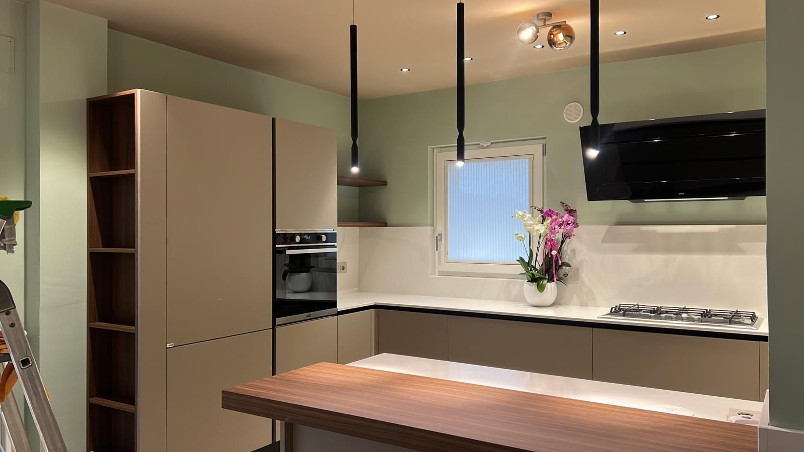 Cucina moderna con mobili beige, isola, lampade a sospensione, forni a incasso e un vaso di fiori vicino alla finestra.
