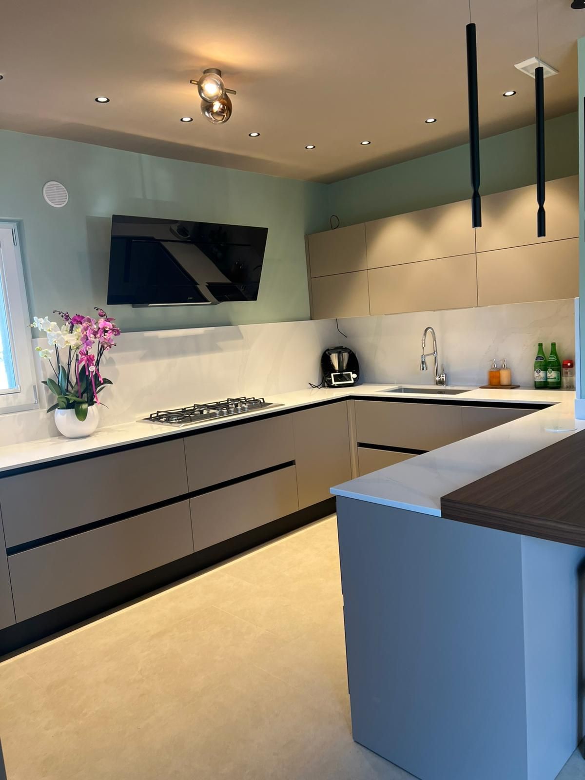 Cucina moderna con mobili chiari, dettagli neri, isola, piano cottura, lavello e lampade a sospensione.
